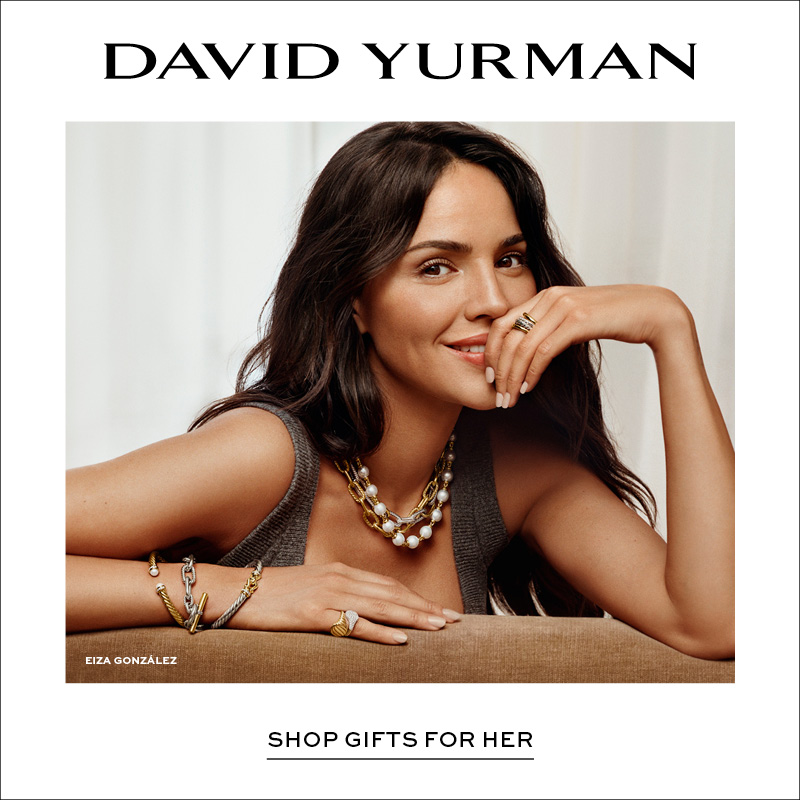 Model wearing layered gold and pearl necklaces, bracelets, and rings, leaning on a neutral-toned sofa with soft drapery background; David Yurman logo and “Shop Gifts for Her” displayed on the right.