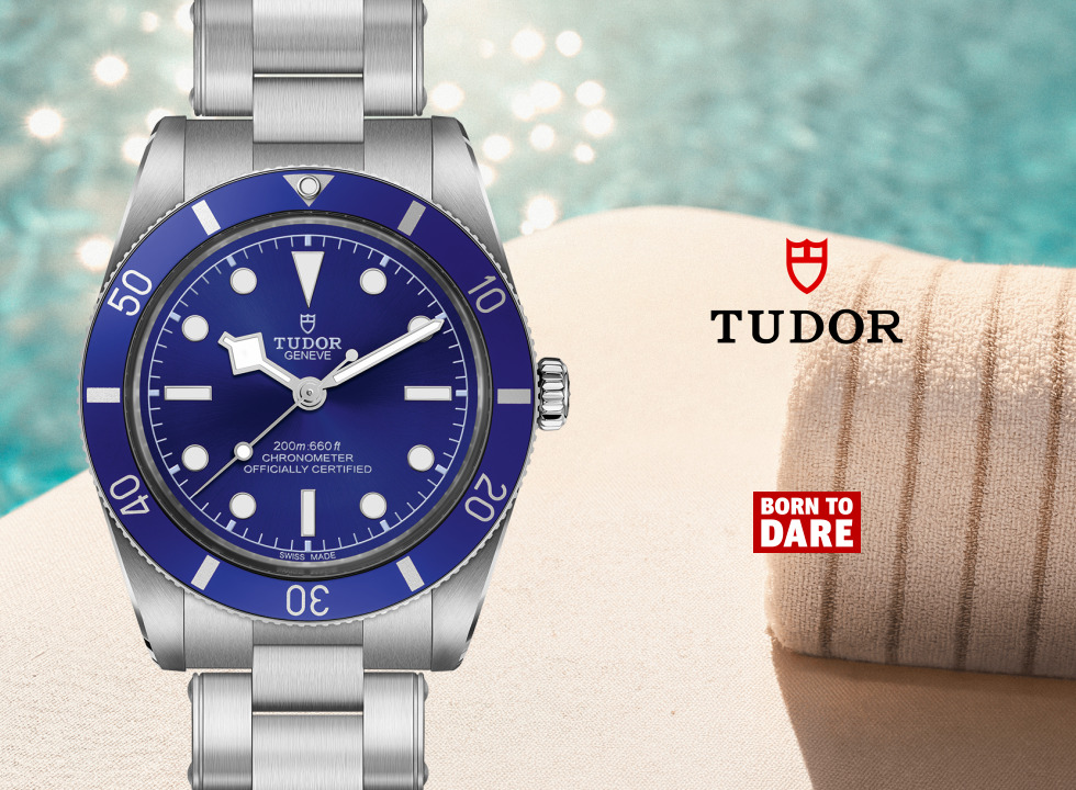 TUDOR Black Bay 54 Blue dive watch with a vibrant blue dial and matching blue bezel on a stainless steel bracelet, displayed beside a rolled beach towel with a sparkling turquoise pool in the background. TUDOR logo and Born To Dare branding appear on the right.